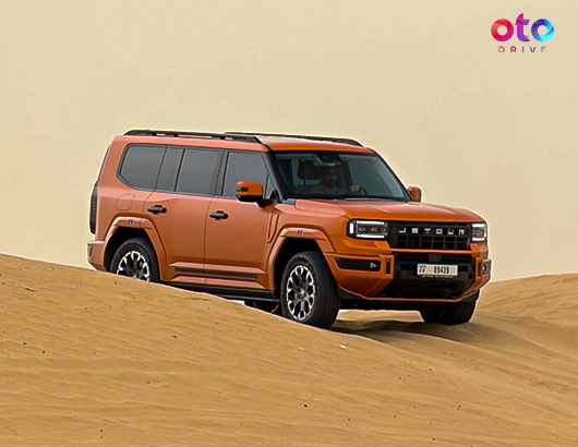 Orange Jetour G700 off-road SUV driving smoothly over desert sand dunes in the sunshine.